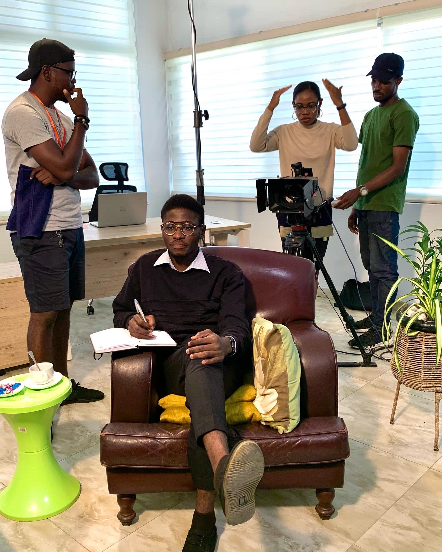 Actor seated in a leather armchair writing in a notebook while crew set up the camera